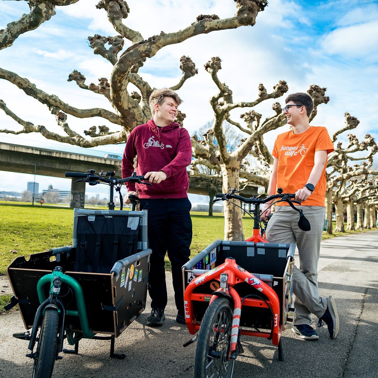 Zwei junge Männer stehen mit Cargobikes auf einem Gehweg, im Hintergrund ist eine Baum-Allee und eine Brücke.