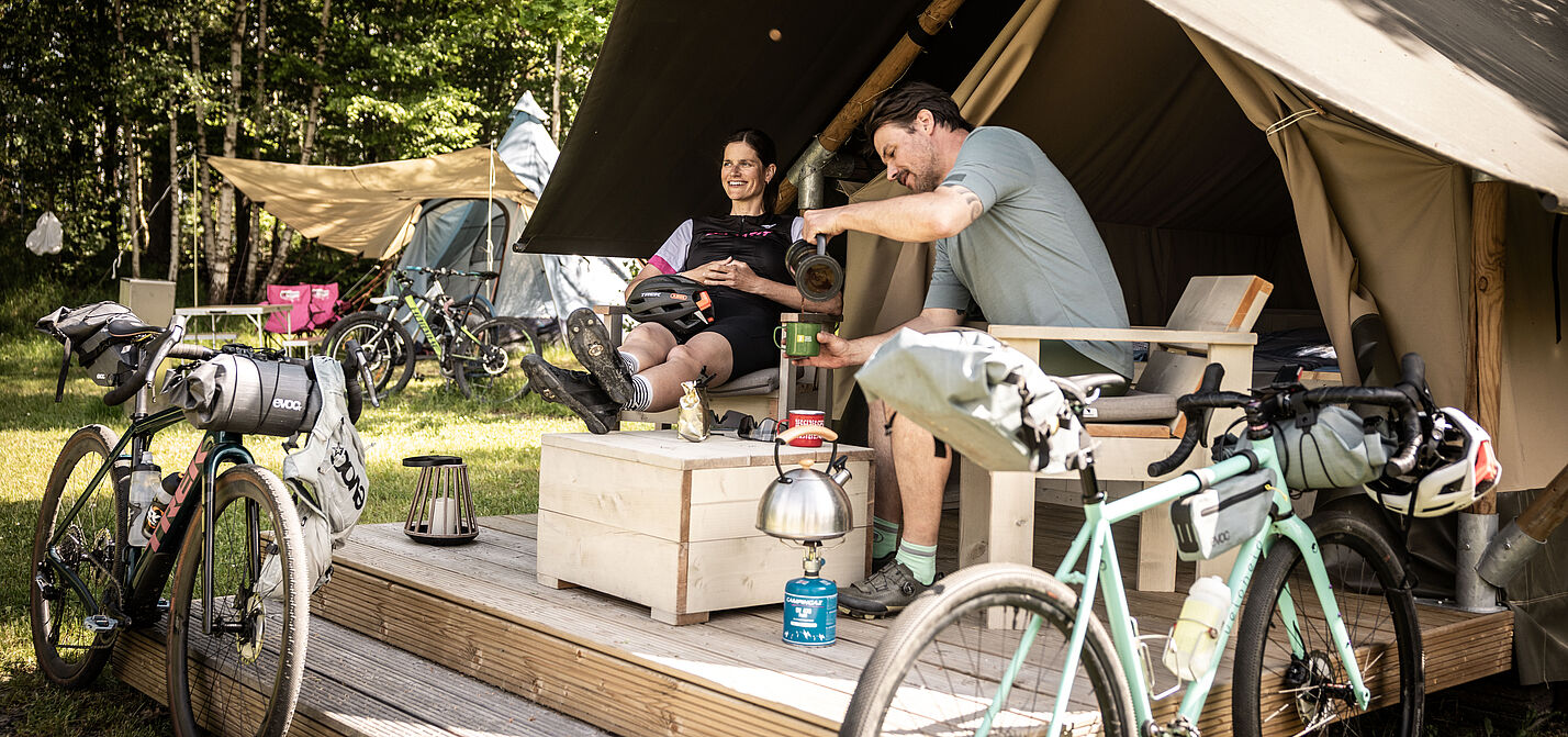 Bett+Bike Sport Unterkunft für Gravelbike Zwei Menschen mit Gravelbikes sitzen im Eingangsbereich auf einer Holzveranda vor einem großen Zelt.