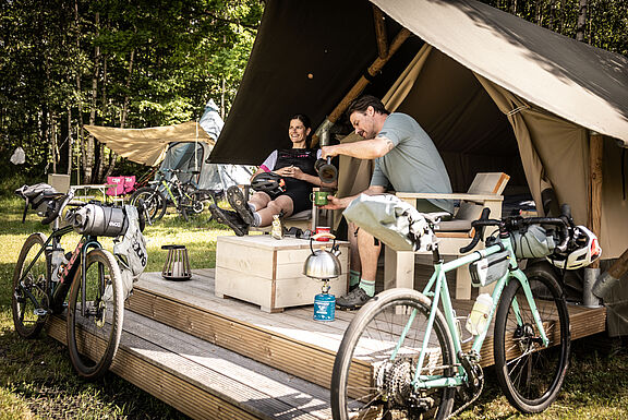Zwei Menschen mit Gravelbikes sitzen im Eingangsbereich auf einer Holzveranda vor einem großen Zelt.
